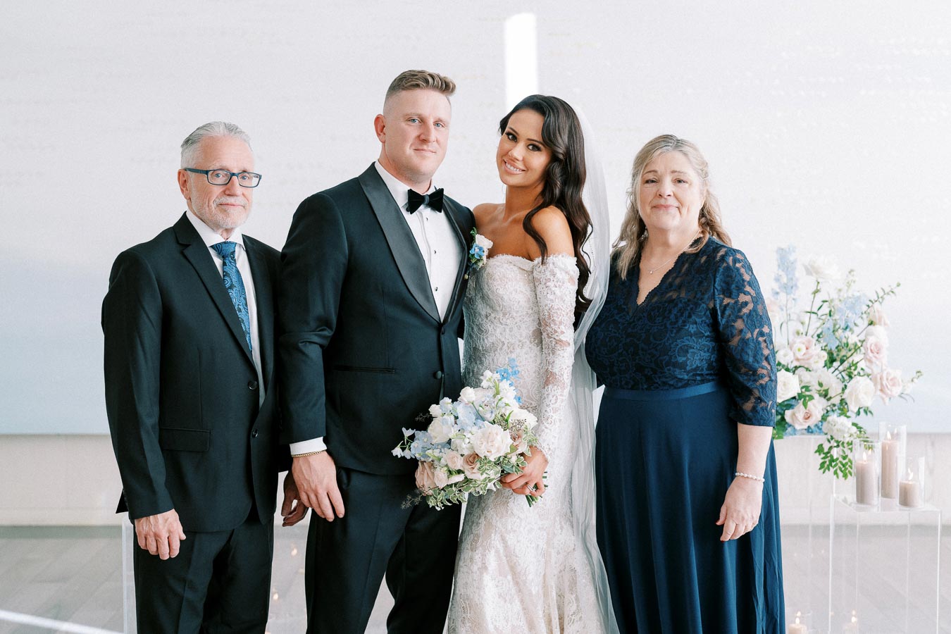 Bride and groom pose with family members in formal attire at an elegant indoor wedding, surrounded by pastel floral arrangements.