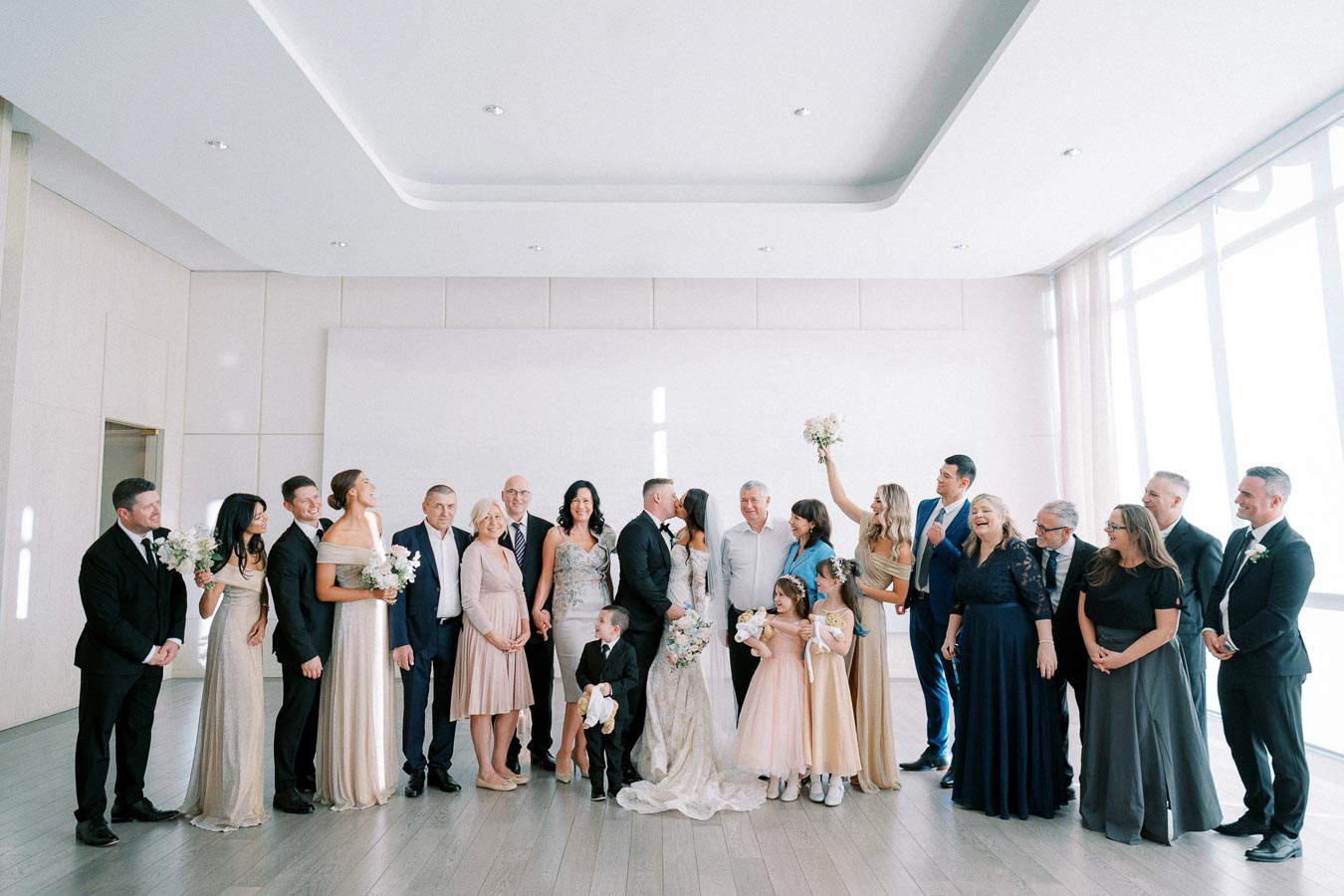 Elegant indoor wedding group photo featuring the bride and groom surrounded by family and friends, dressed in formal attire, celebrating together in a bright, spacious room.