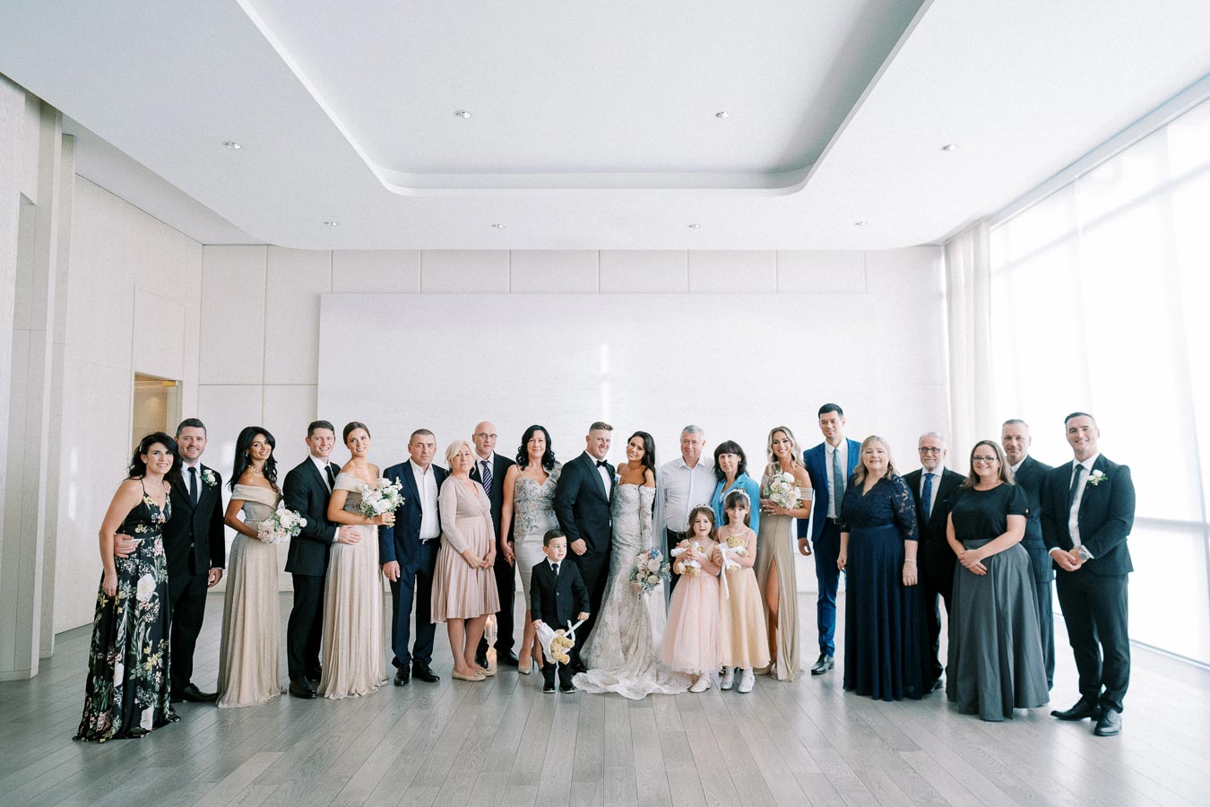 A large group photo of a wedding party in a bright room, featuring the bride and groom surrounded by family and friends. The group is dressed in formal attire, with bridesmaids in elegant dresses holding bouquets and groomsmen in suits. The room has modern decor with natural light streaming in from large windows.