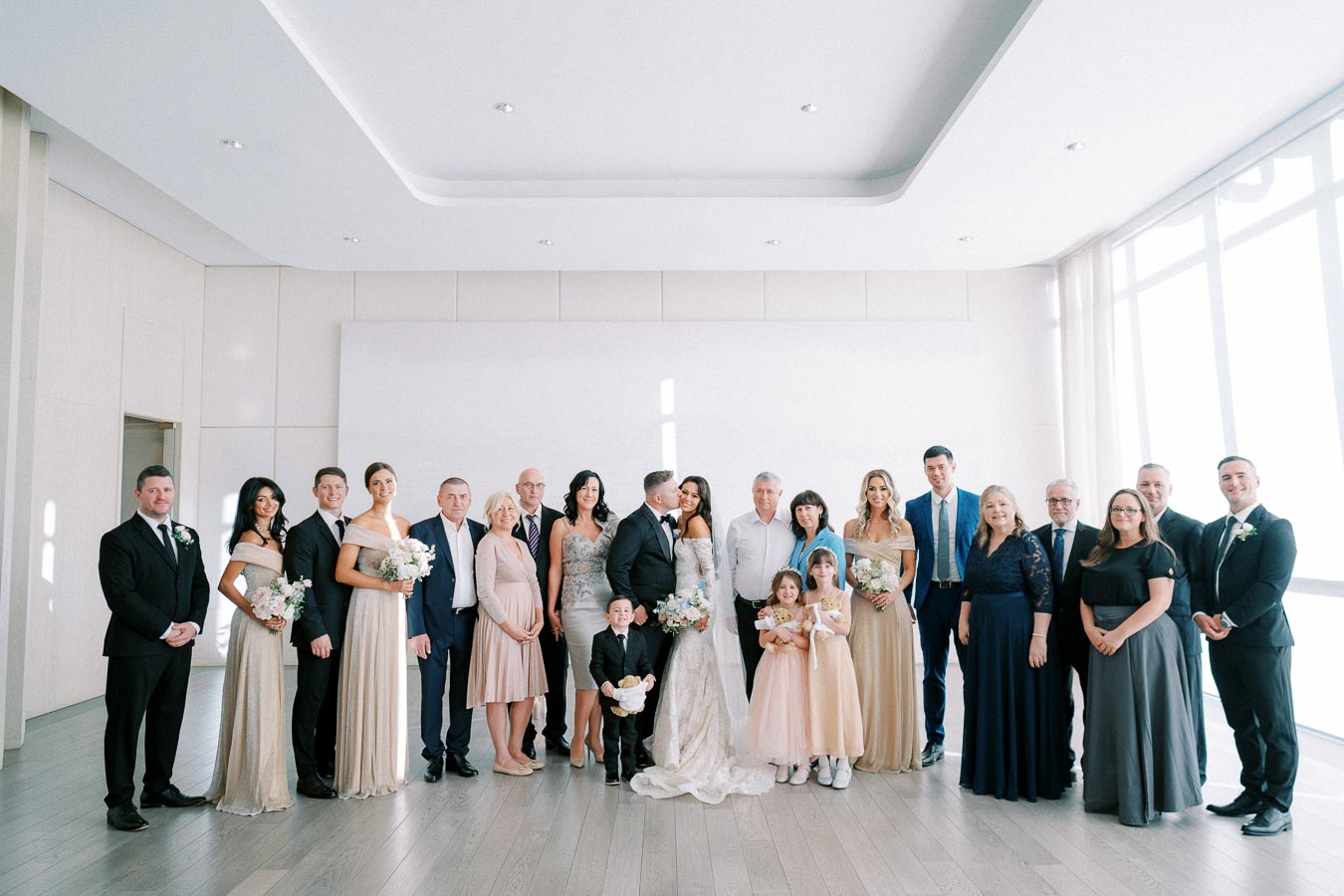 Wedding group photo in a bright room, featuring a diverse wedding party and family members dressed in formal attire, with the bride and groom in the center. The bride is wearing a white dress and the groom a black suit, surrounded by bridesmaids, groomsmen, and flower girls holding bouquets.