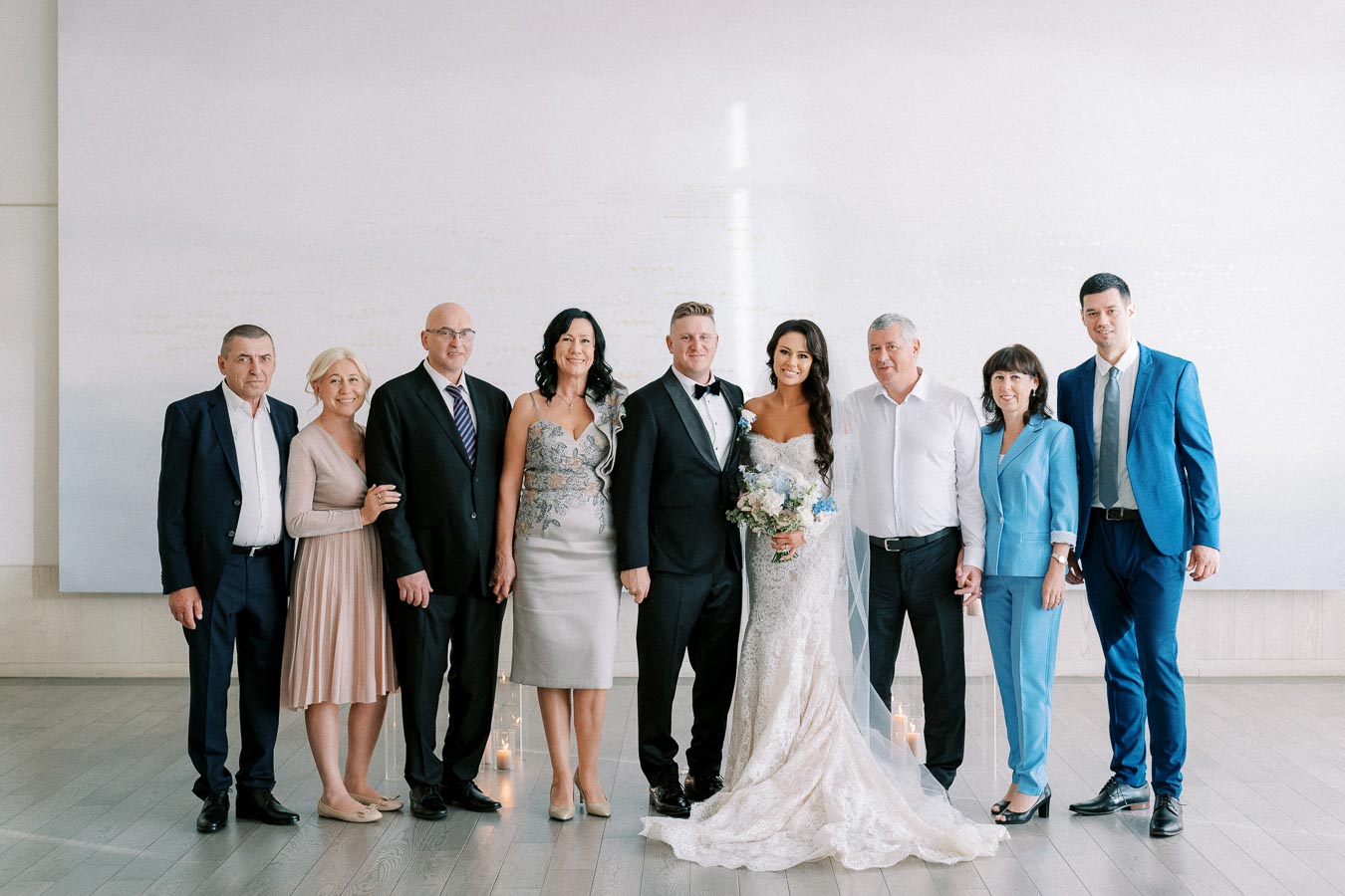 Group photo of a bride and groom in formal attire with family members in elegant dresses and suits, celebrating a wedding indoors.