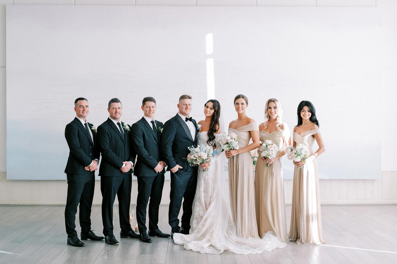 A wedding party group photo featuring the bride in a white gown and six attendants in black suits and gold dresses, posing indoors with bouquets of flowers.