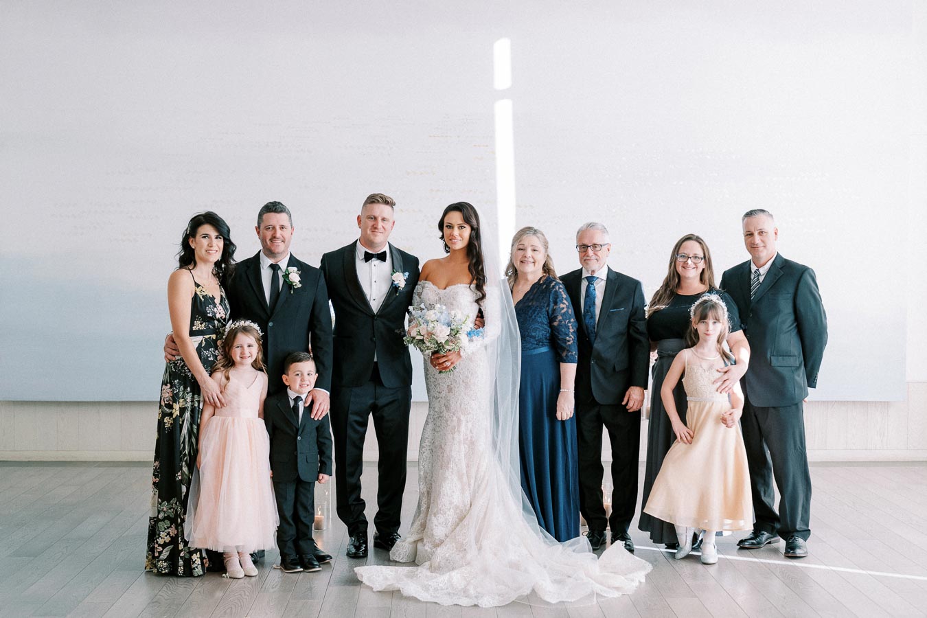 A joyful wedding portrait featuring a bride in an elegant lace gown and groom in a black suit, surrounded by family members dressed in formal attire, all smiling against a simple background.