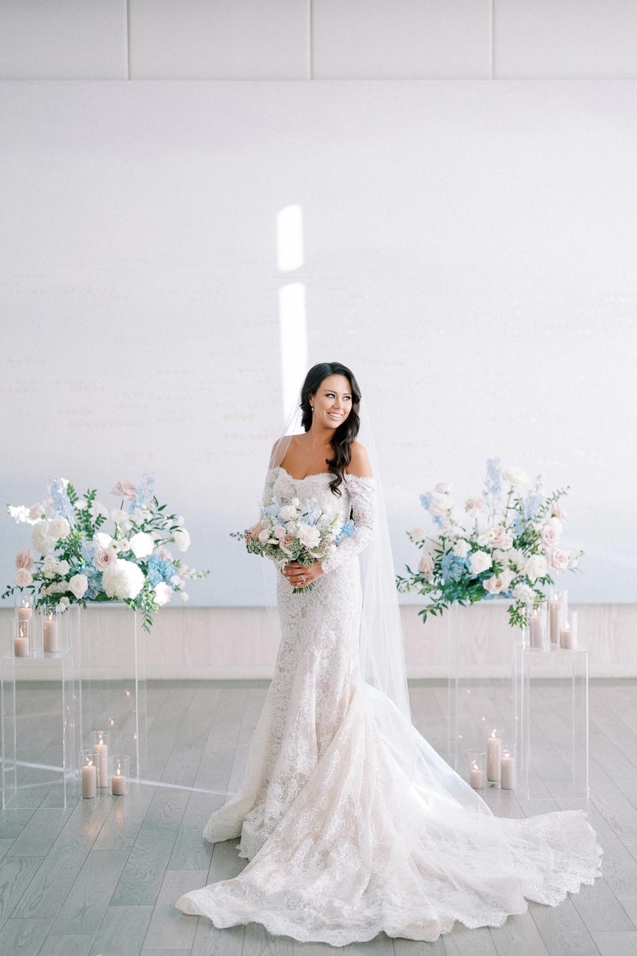 Beautiful bride in lace wedding gown holding a bouquet of pastel flowers, standing between elegant floral arrangements and candles in a bright, minimalist venue.