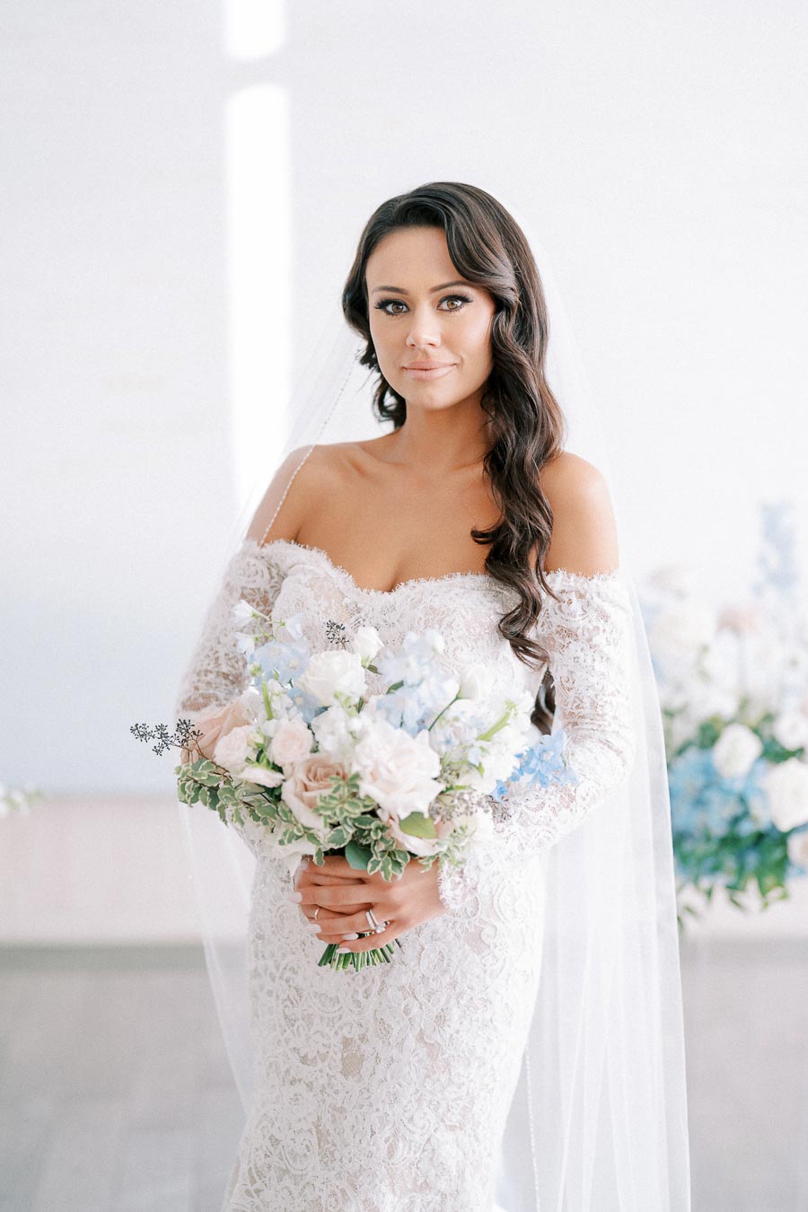 Elegant bride in lace wedding dress holding pastel bouquet, standing indoors with soft natural lighting.
