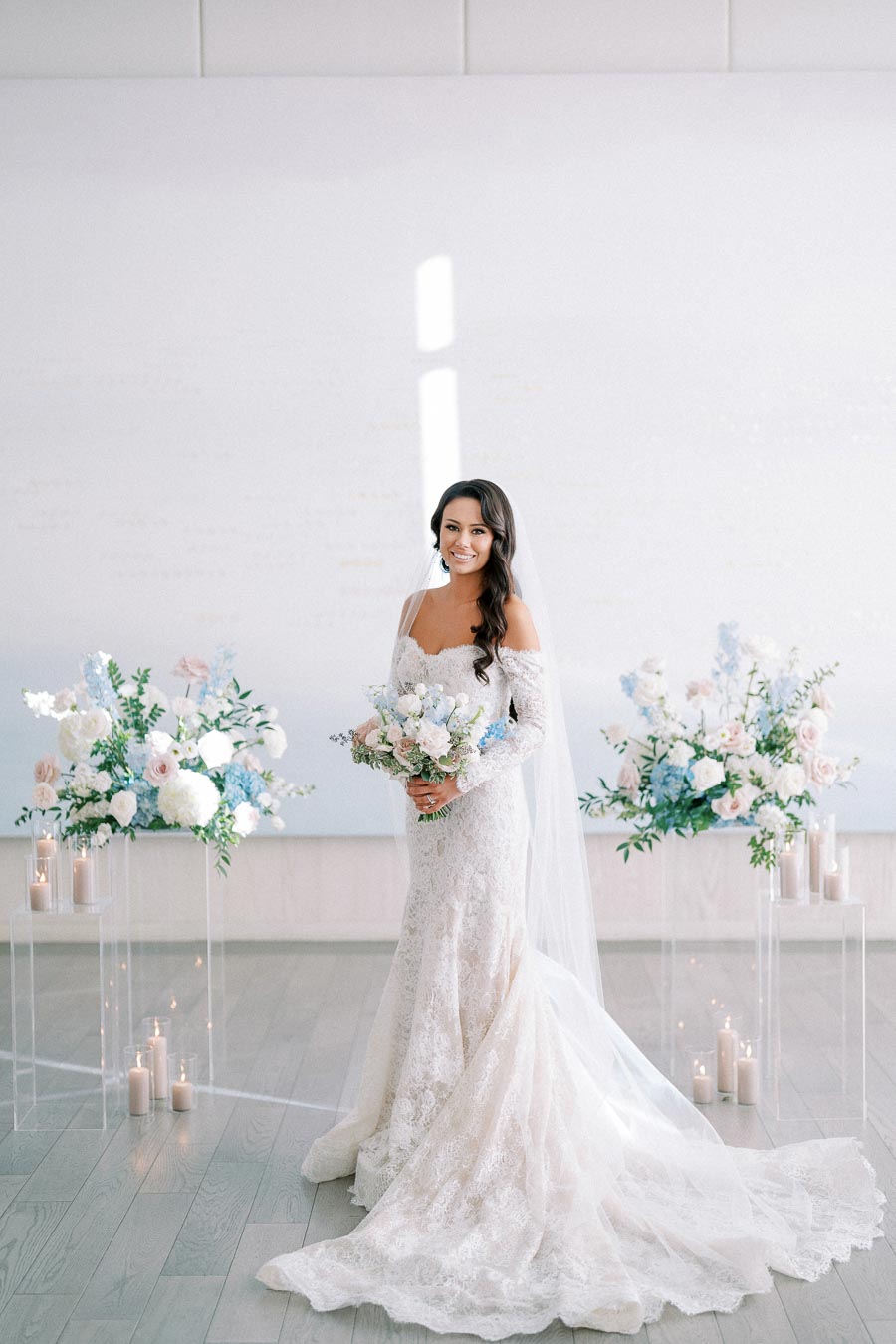 Bride in an elegant lace wedding dress holding a bouquet, surrounded by floral arrangements and candles in a bright, minimalist venue.