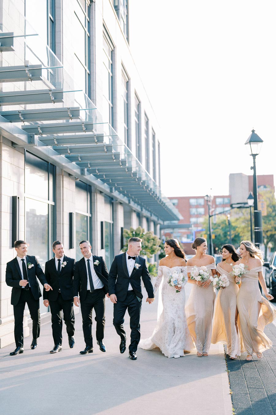 Wedding party walking outside a modern building, featuring a bride in a lace gown holding flowers, bridesmaids in matching dresses, and groomsmen in black suits with boutonnieres.