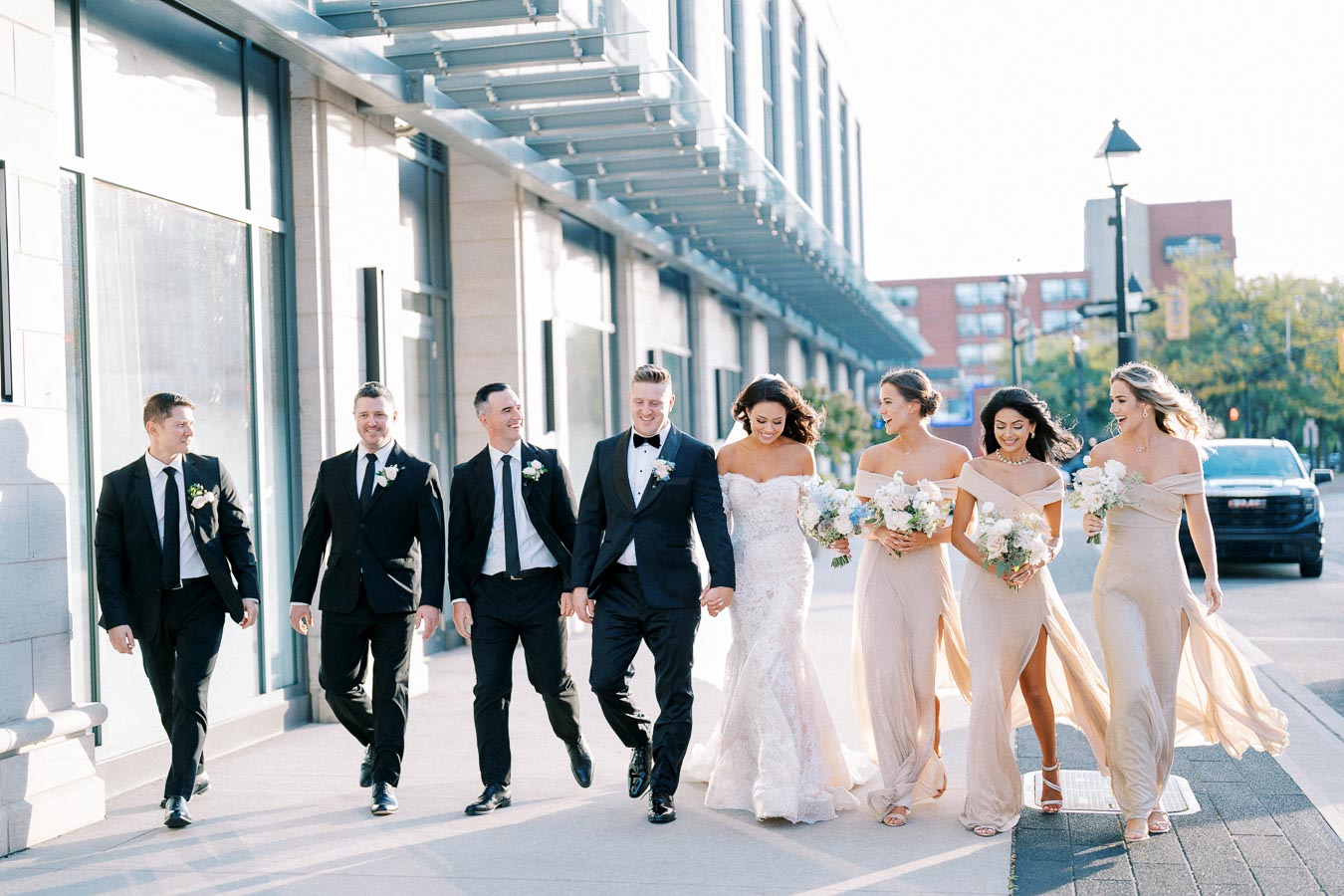 A joyful bridal party walks down a city street, with the bride in a white gown and bridesmaids in matching beige dresses, alongside groomsmen in black suits, under a sunny blue sky.