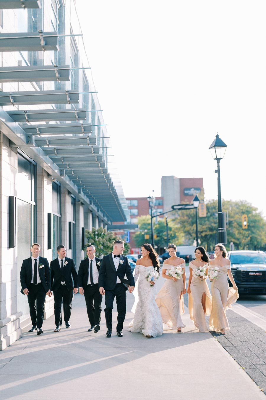 A bride and groom walk with bridesmaids and groomsmen on a sunny urban street, captured in elegant wedding attire.