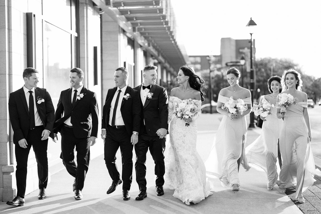 A joyful bridal party walking outdoors, featuring the bride in a lace gown and holding flowers, accompanied by bridesmaids in matching dresses and groomsmen in suits.