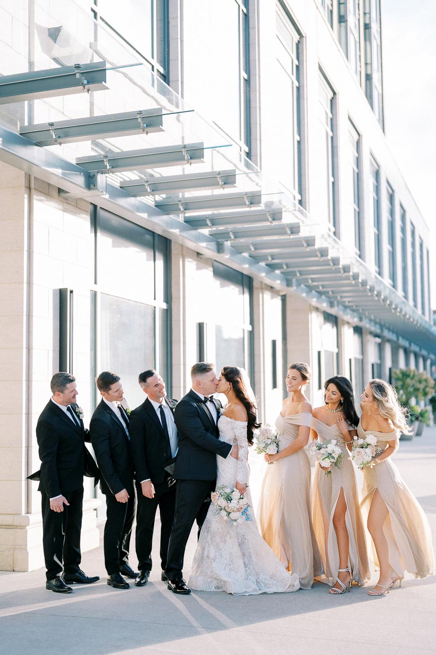 Wedding party posing outside a modern building, featuring a bride in an elegant lace gown kissing the groom, surrounded by bridesmaids in light champagne dresses and groomsmen in black suits, all holding floral bouquets.