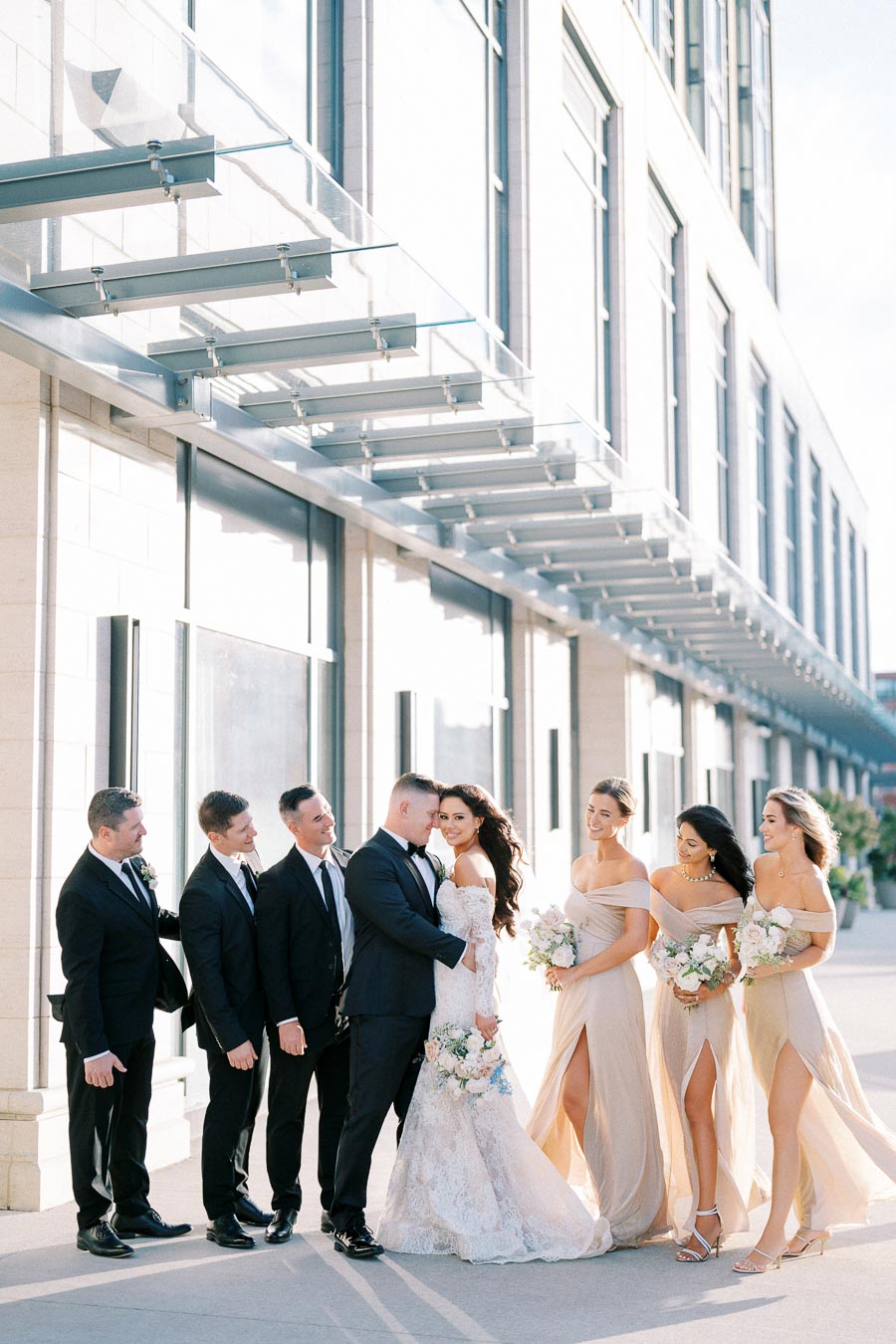 A wedding party standing outdoors, featuring a bride in a lace gown and groom in a black suit, surrounded by bridesmaids in beige dresses and groomsmen in black suits, posing against a modern building background.