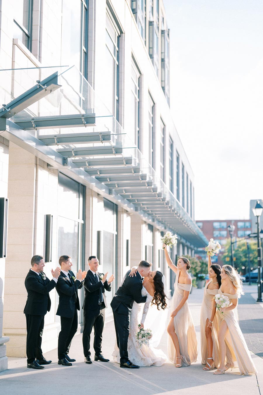 Newlywed couple kissing outside, surrounded by groomsmen in suits and bridesmaids in champagne dresses, celebrating under a glass awning of a modern building.