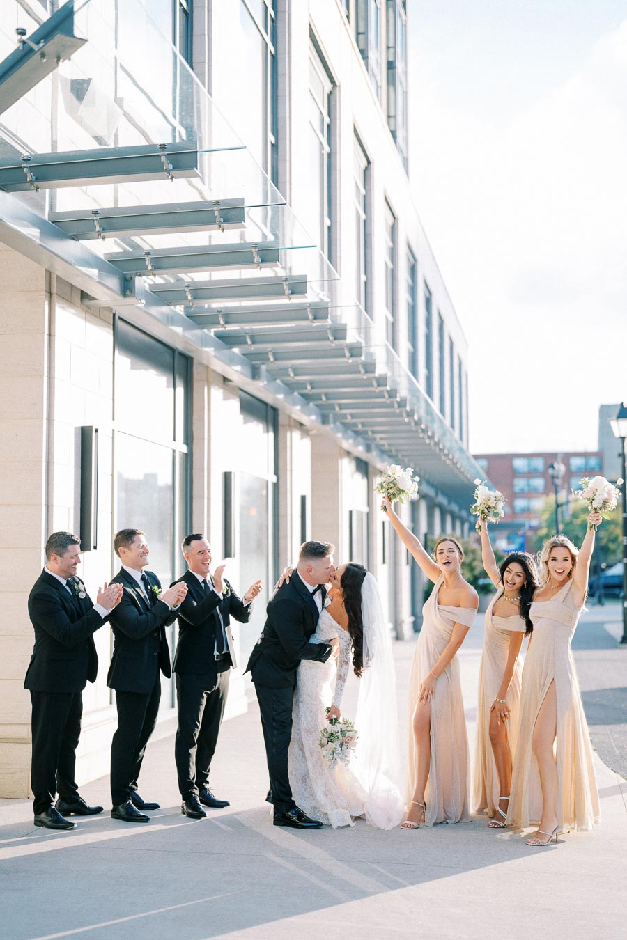 Bride and groom kiss outside modern building, surrounded by groomsmen in suits clapping and bridesmaids in matching neutral dresses holding bouquets and celebrating in sunlight.