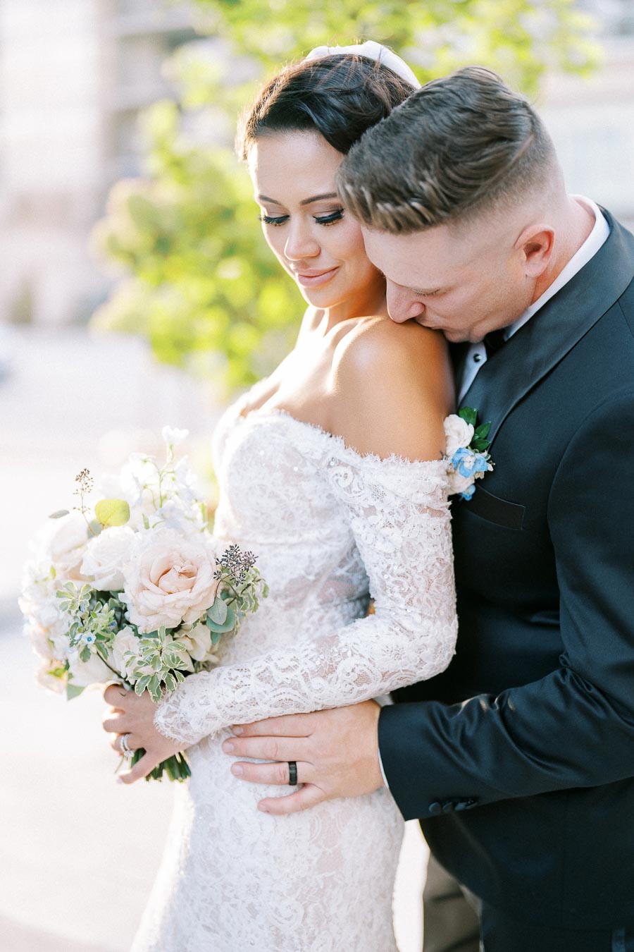 Elegant bride in a lace wedding dress holding a bouquet, embraced by her groom in a tender moment outdoors.