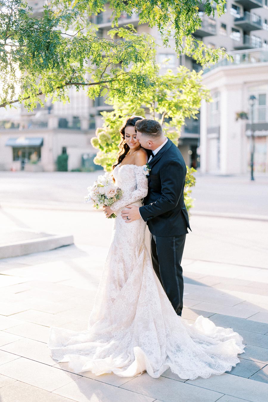 A bride in an elegant lace wedding dress and a groom in a black suit embrace tenderly under a tree, with an urban backdrop and soft sunlight illuminating the scene.