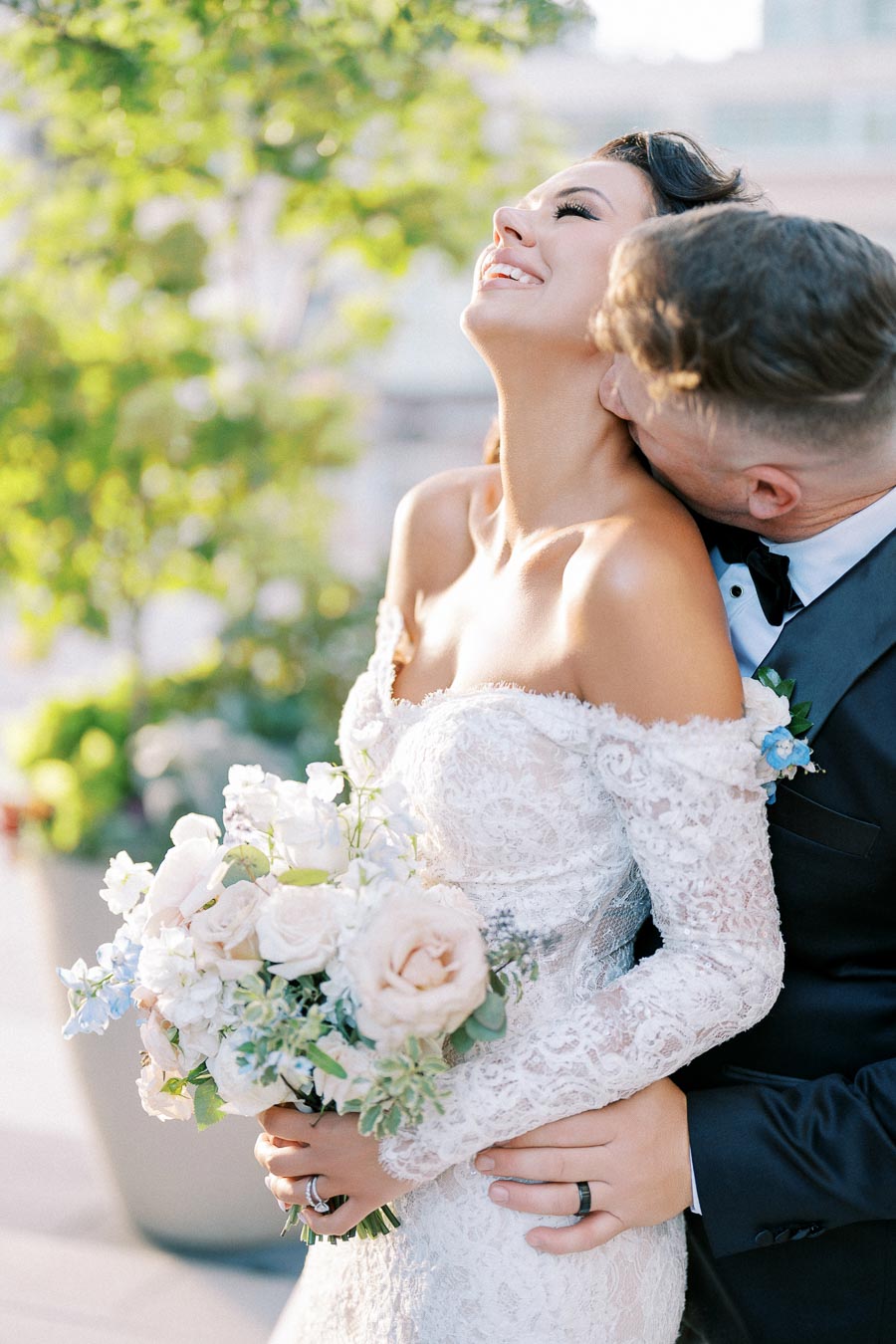Bride in lace wedding gown joyfully embraces groom with bouquet of pastel flowers, outdoors in lush garden setting.