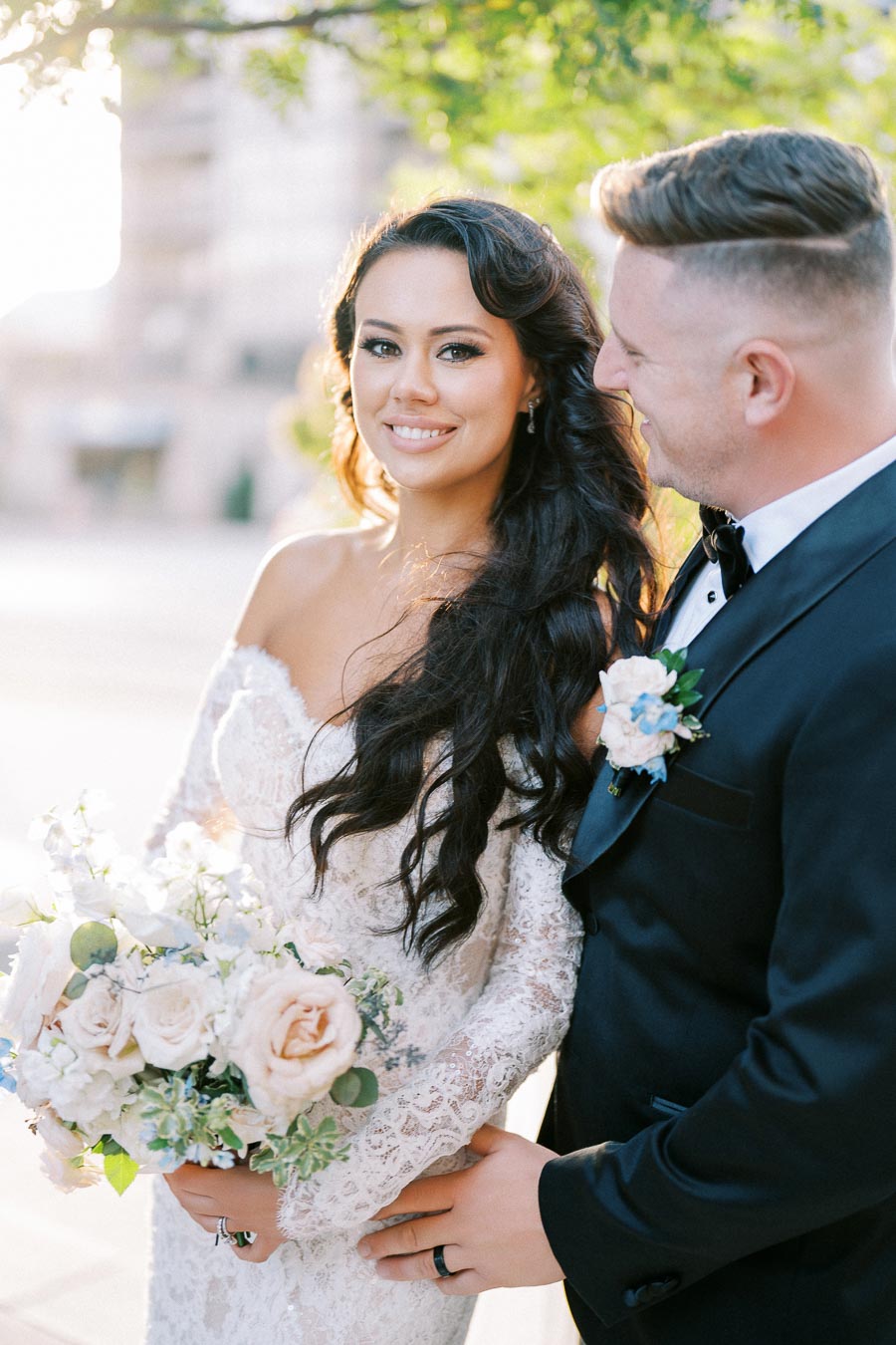Elegant bride in lace wedding dress holding bouquet, standing with groom in tuxedo, outdoor wedding photo with soft sunlight and greenery.