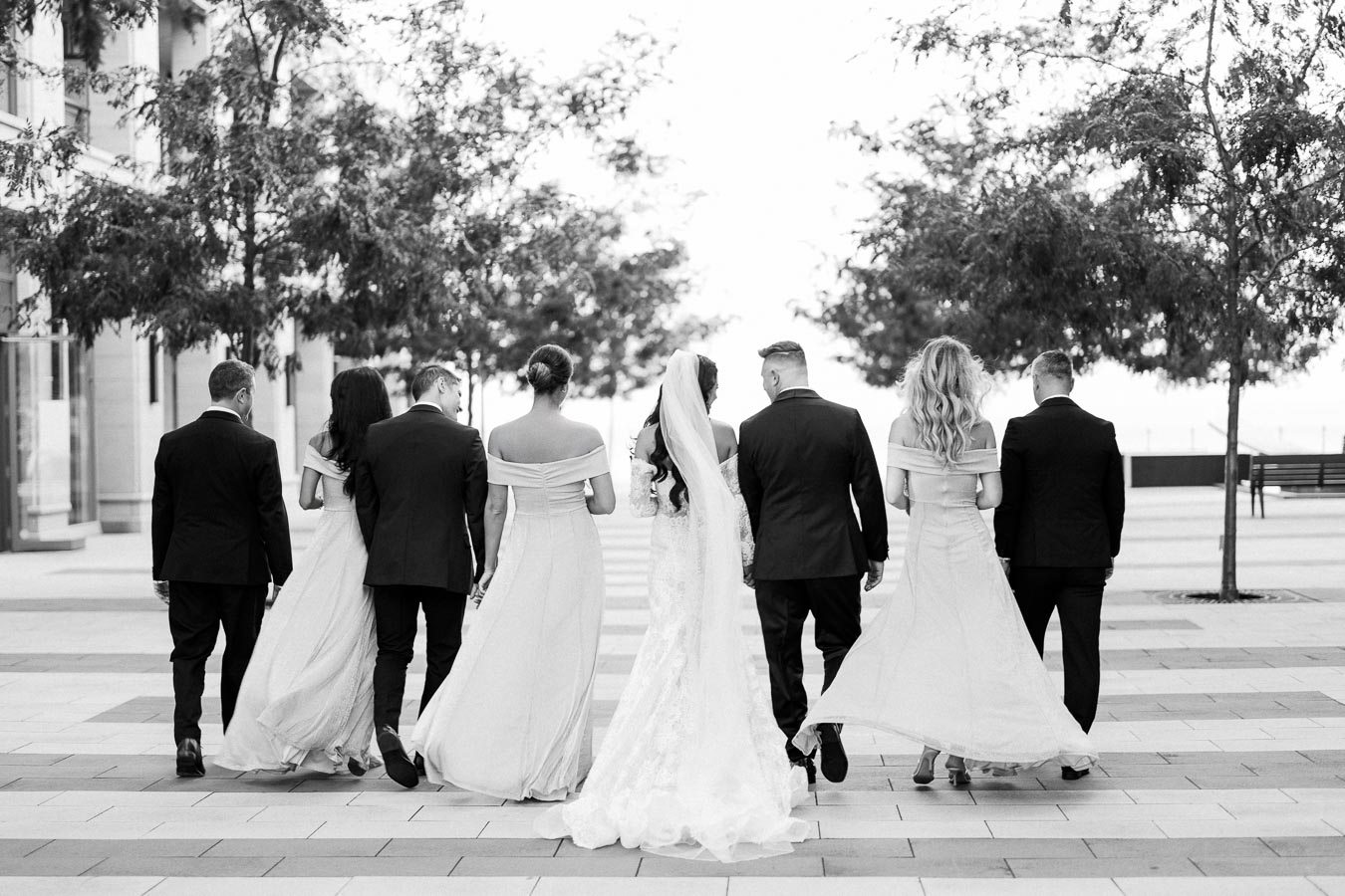 Black and white photo of a wedding party walking away, featuring a bride in a long gown with veil and bridesmaids in matching dresses, accompanied by groomsmen in suits, against an urban backdrop with trees.