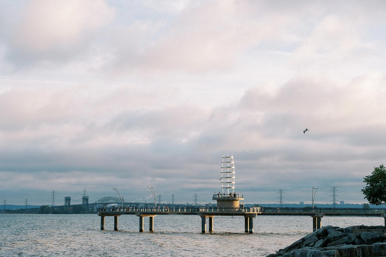 Scenic view of Burlington Pier extending over Lake Ontario under a cloudy sky, with distant bridges and industrial structures in the background, and a seagull flying overhead.