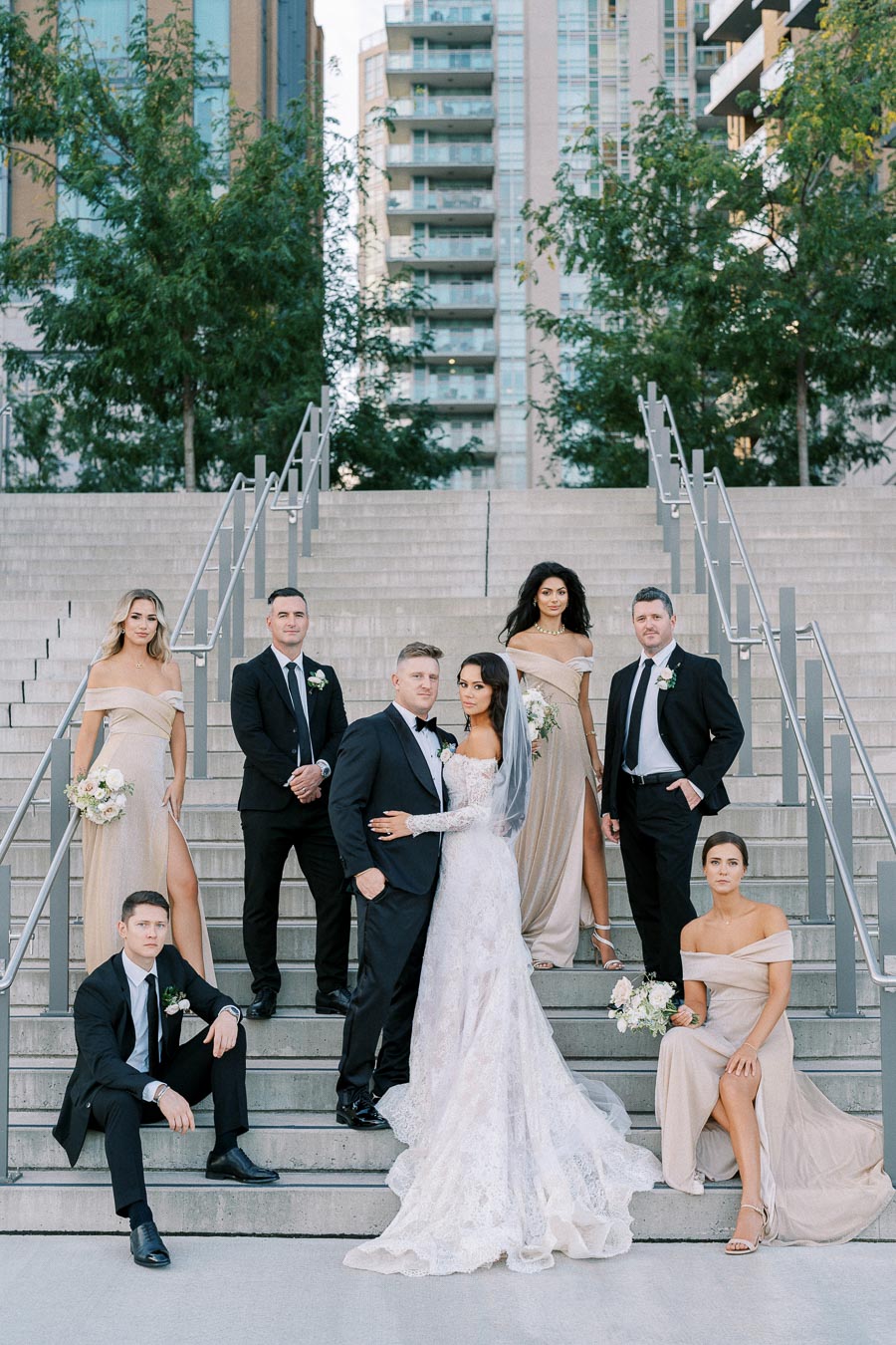 Elegant wedding party posing on modern city steps, bride in lace gown with veil, surrounded by bridesmaids in taupe dresses and groomsmen in black suits, against an urban backdrop with high-rise buildings and greenery.