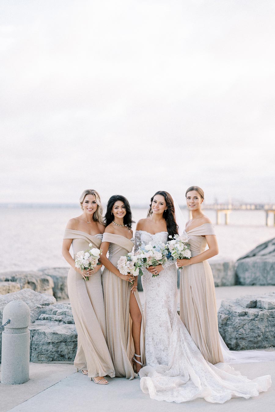 Bride and bridesmaids in elegant gowns pose with bouquets by the waterfront, with a scenic view of the ocean and a bridge in the background.