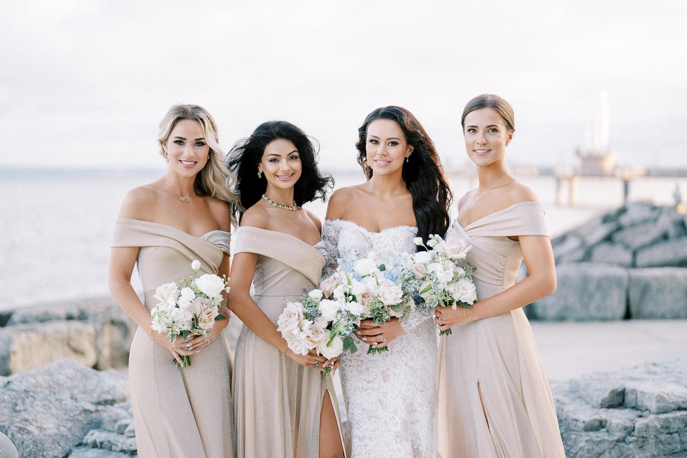 Four women in elegant bridesmaid dresses stand together holding floral bouquets, with a scenic rocky waterfront in the background.