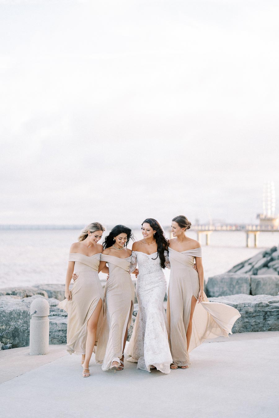 Bridal party smiling and walking along a scenic waterfront, showcasing elegant off-shoulder beige bridesmaid dresses and a lace wedding gown, with a pier and ocean in the background.