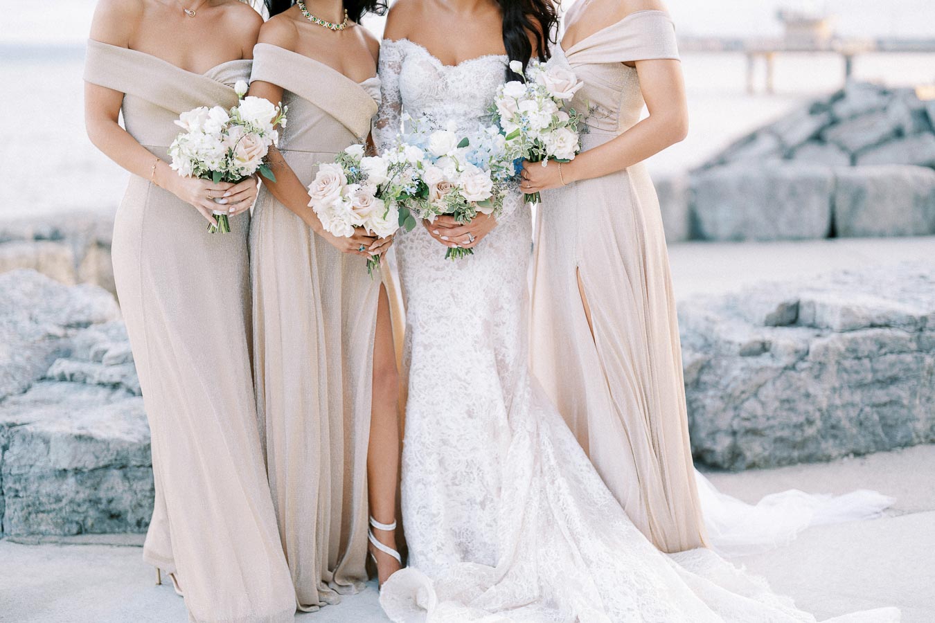 Bridal party at seaside wedding, featuring the bride in a lace gown with bridesmaids in elegant beige dresses, holding pastel floral bouquets against a rocky ocean backdrop.