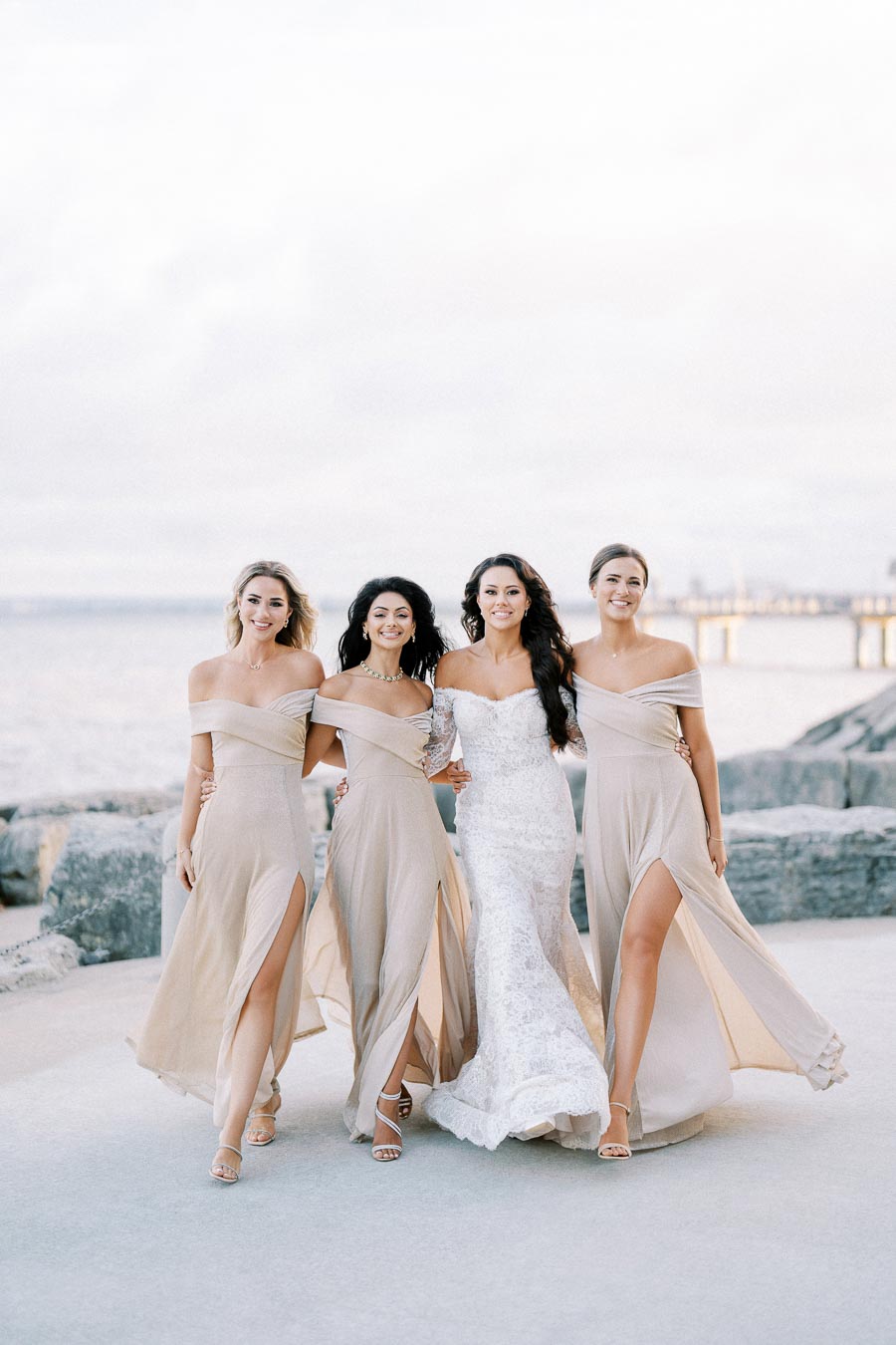 Bridesmaids in elegant pale dresses walking with the bride in a white lace gown along a scenic coastal path.