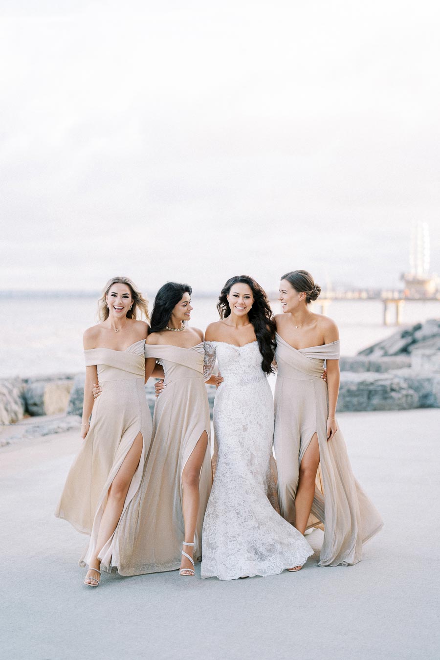 A bride in a white lace gown walks arm-in-arm with her three bridesmaids in matching beige dresses with slits, set against a scenic waterfront backdrop.