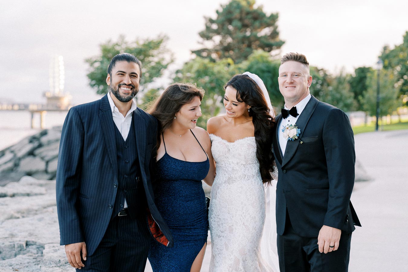 A joyful wedding moment by the lake with a bride in a lace gown, groom in a tuxedo, and two elegantly dressed guests sharing smiles and laughter.