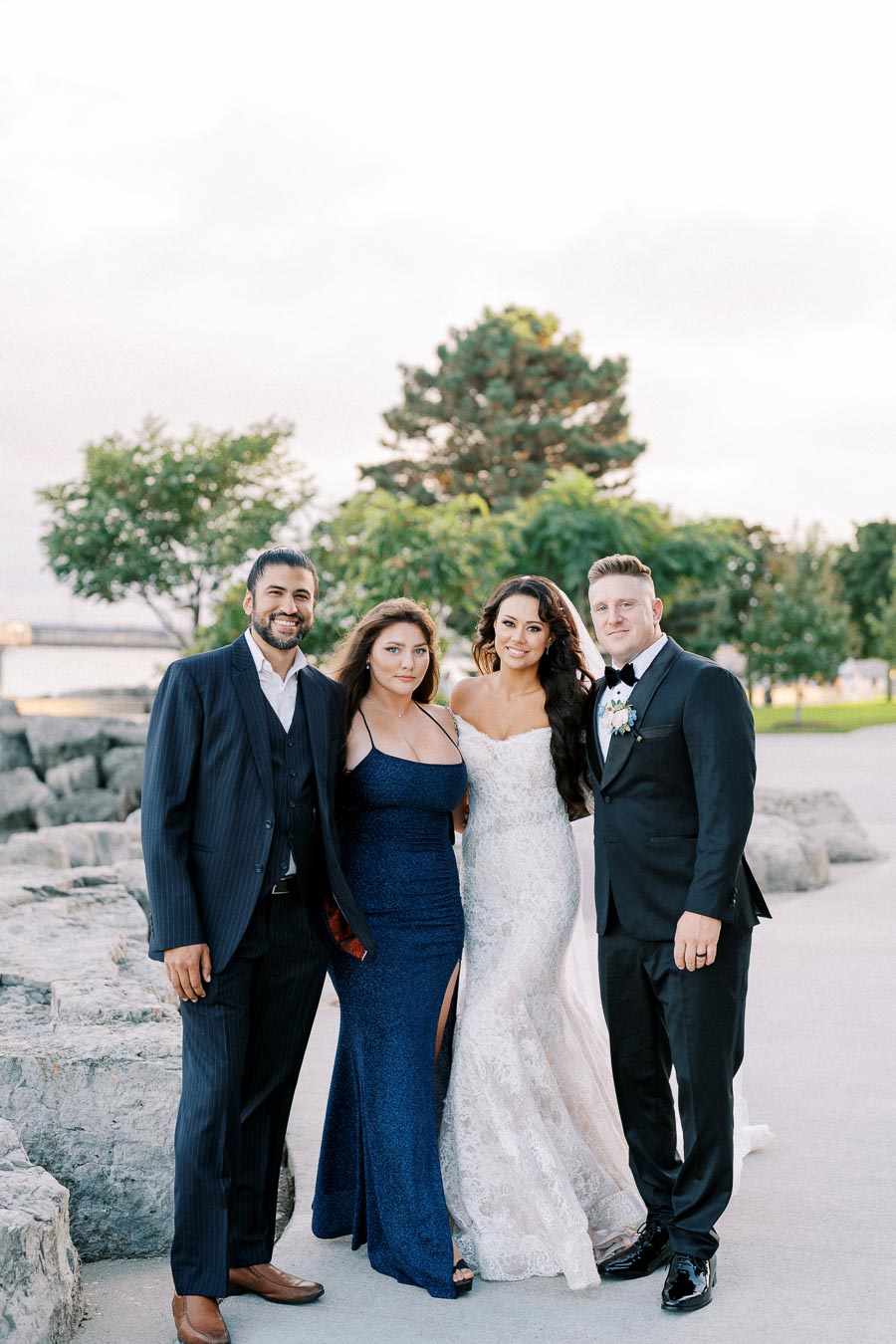 A group of four people dressed elegantly pose together outdoors, featuring two men in suits and two women in formal dresses, with a scenic background of trees and rocks.
