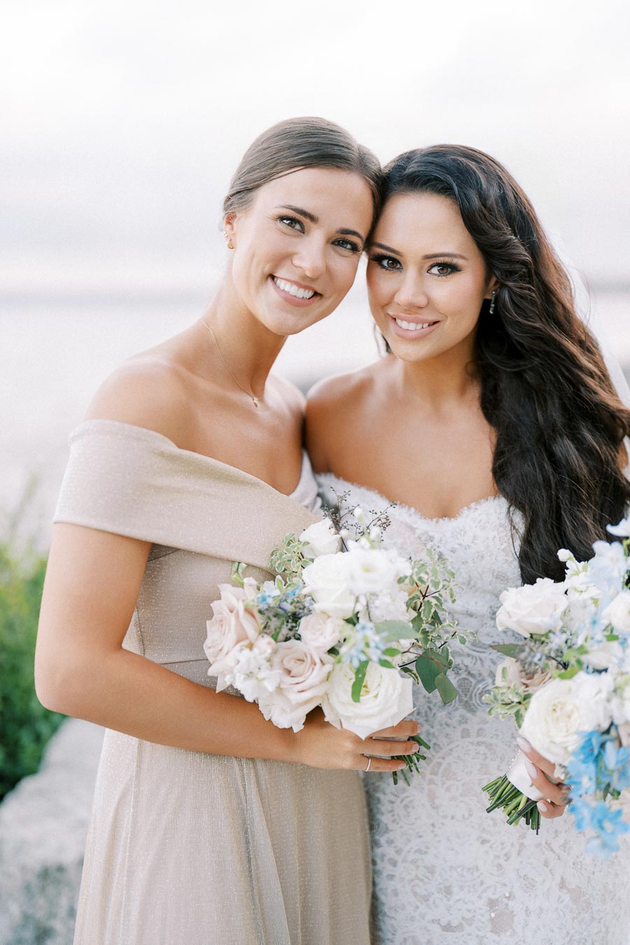 Two women in elegant gowns holding bouquets of flowers at a wedding, smiling with a serene outdoor backdrop.