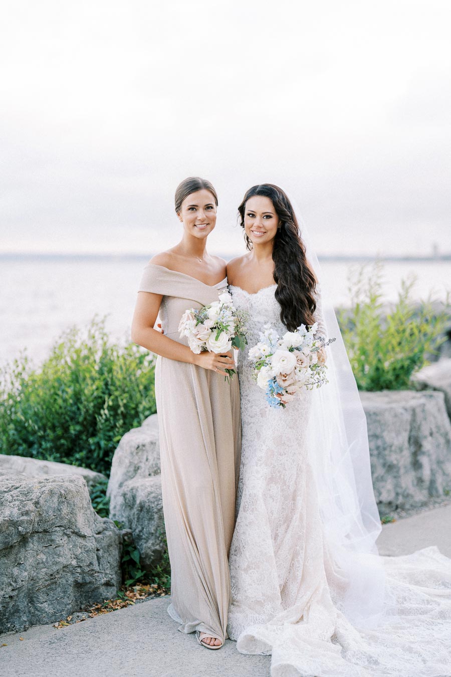 Two women in elegant dresses pose by a rocky lakeside. The woman on the right is wearing a white lace wedding gown, holding a bouquet of white and blue flowers. The woman on the left is in a beige off-shoulder dress, holding a matching bouquet. The natural backdrop enhances the serene, picturesque wedding setting.