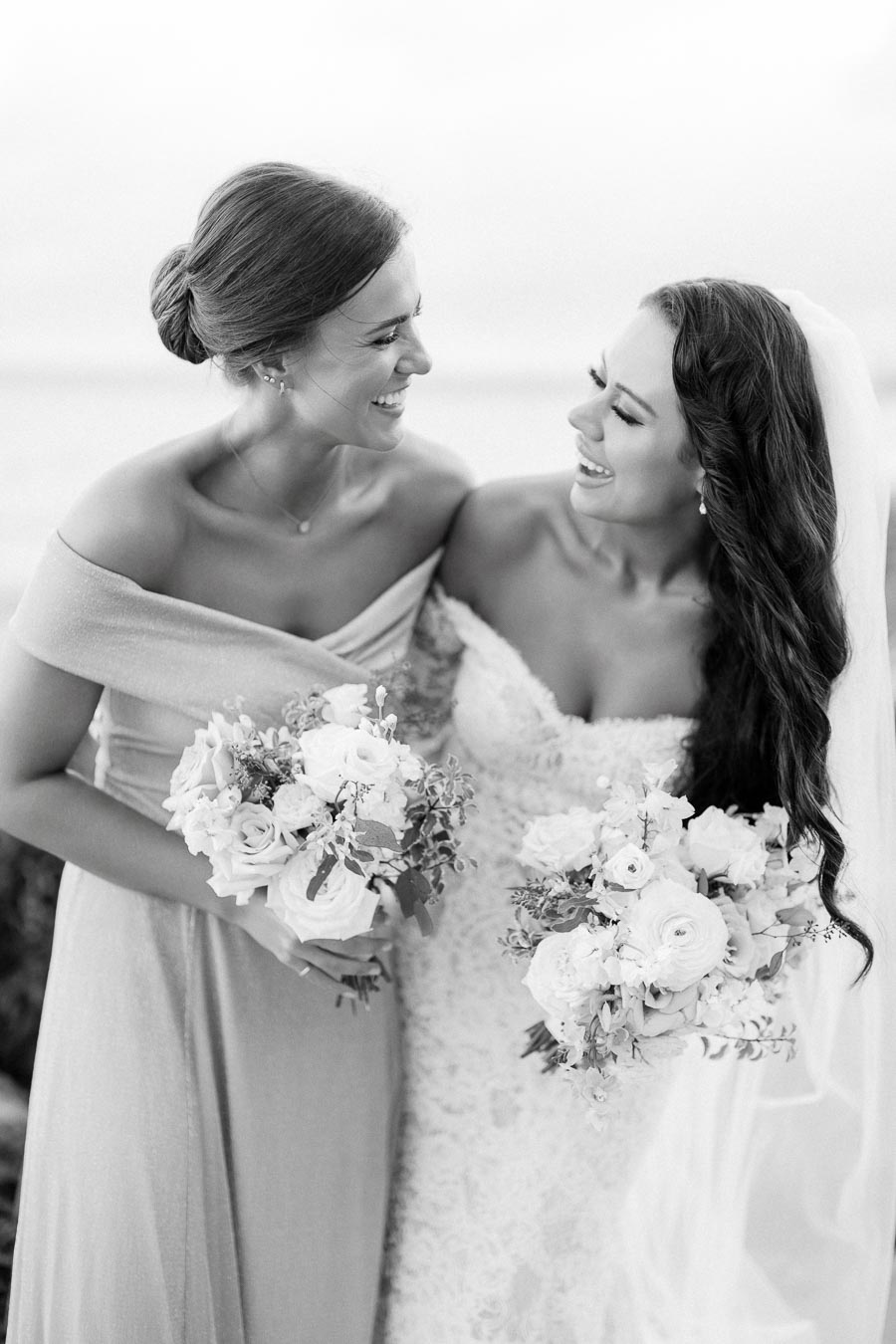 Black and white photo of a smiling bride and bridesmaid holding floral bouquets, wearing elegant dresses with the ocean in the background.
