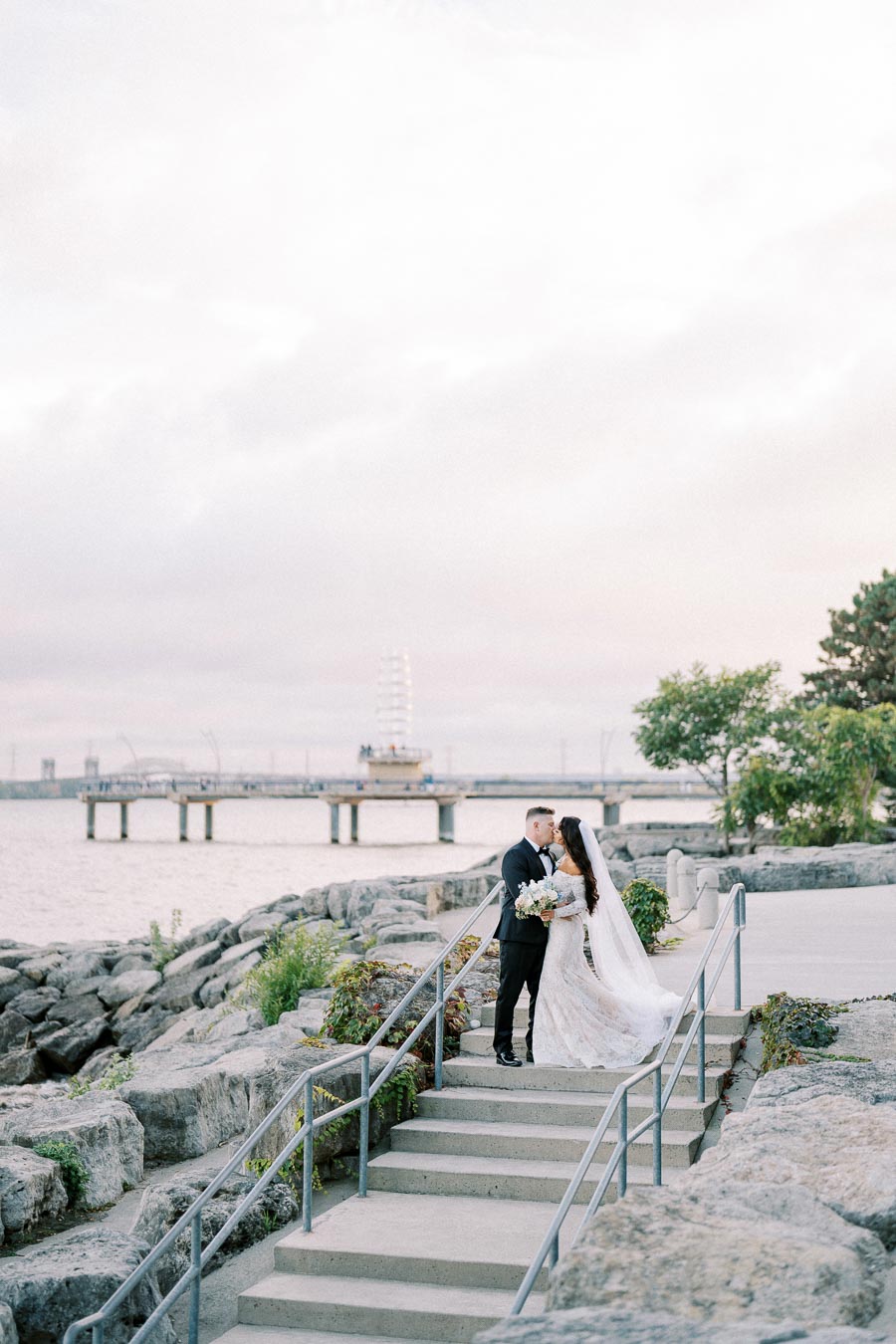 A bride and groom embrace on stone steps by the waterfront, with a scenic pier and cloudy sky in the background.