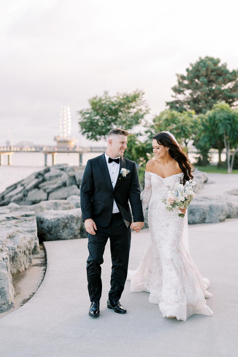 A bride and groom walking hand in hand along a scenic waterfront path, with the bride in a lace off-shoulder gown holding a bouquet, and the groom in a black tuxedo, surrounded by trees and rock formations.