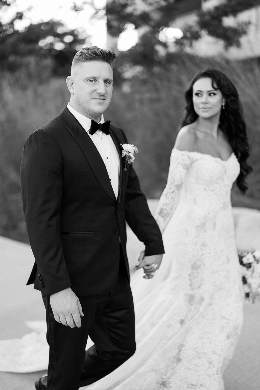 A black and white image of a bride and groom holding hands. The groom is wearing a classic black tuxedo with a bow tie, and the bride is in an elegant lace wedding dress. They are outdoors, walking together with soft focus greenery in the background.