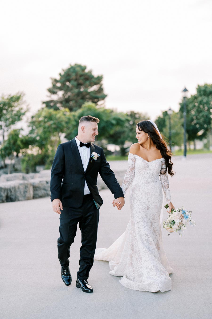 Newlywed couple holding hands and smiling during a romantic outdoor photoshoot, with the bride in an elegant off-shoulder lace wedding gown and the groom in a classic black tuxedo, surrounded by lush greenery.