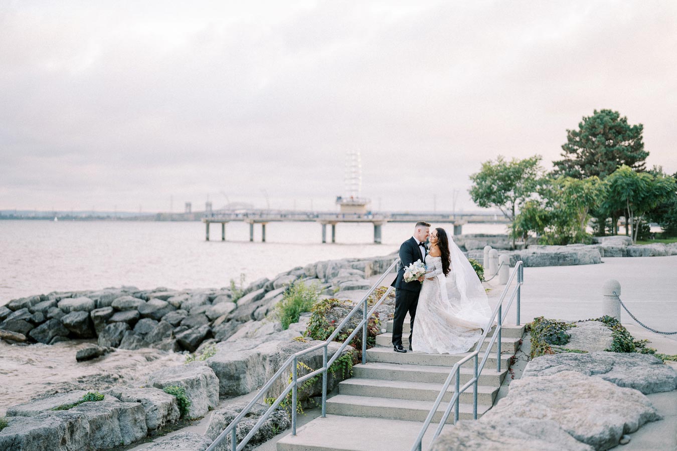 A newlywed couple embracing on a stone staircase by a waterfront, with a scenic view of a pier and cloudy sky in the background. Bride wearing a flowing white gown with a veil, holding a bouquet, while the groom is in a dark suit.