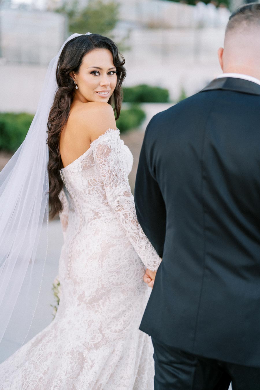 Beautiful bride in a lace wedding dress and veil smiling while holding hands with groom in black suit.