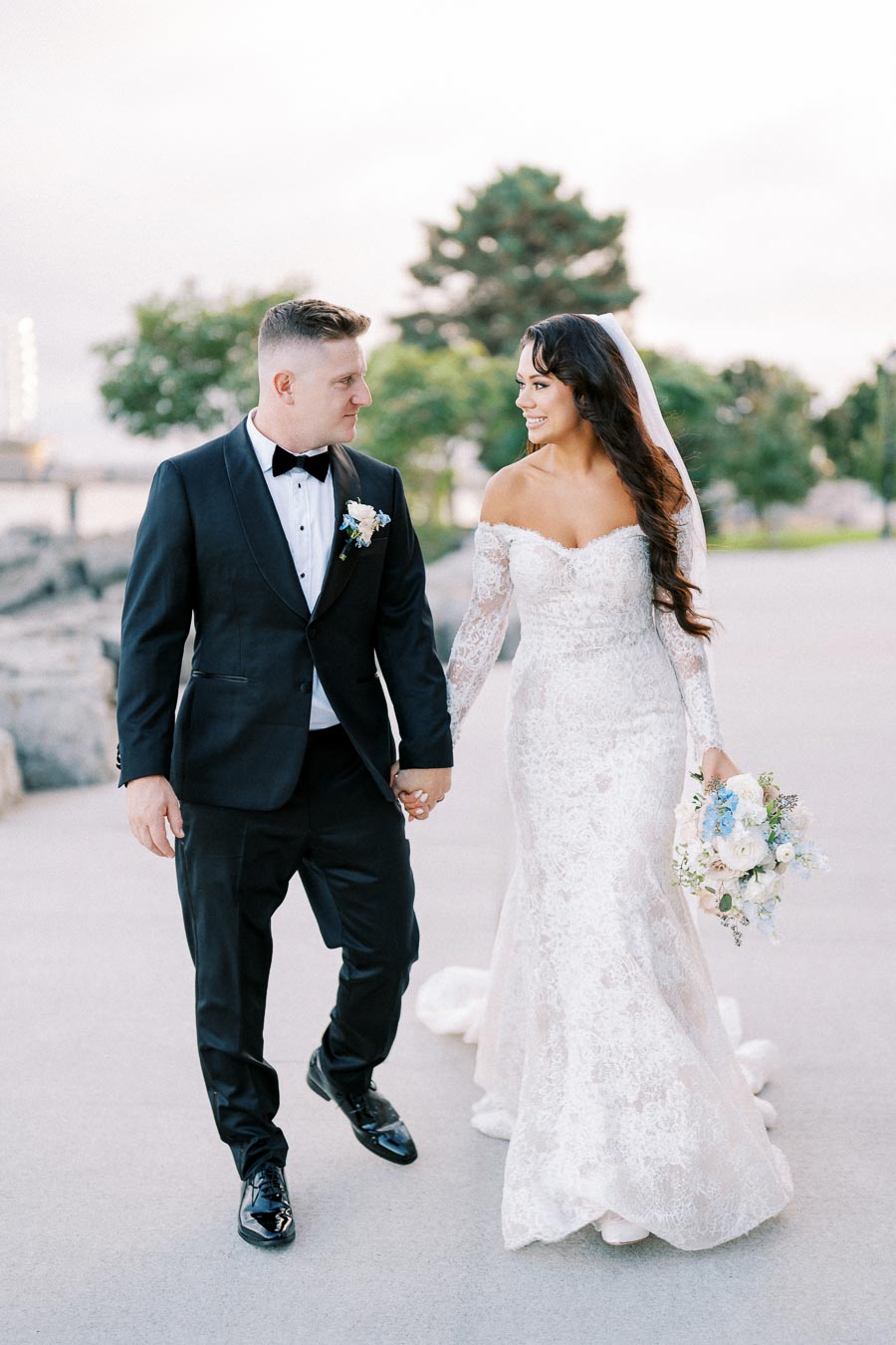 A bride and groom walking hand in hand in elegant wedding attire; the bride in a lace off-shoulder gown holding a bouquet with white and blue flowers, and the groom in a black tuxedo, set against a serene outdoor backdrop.