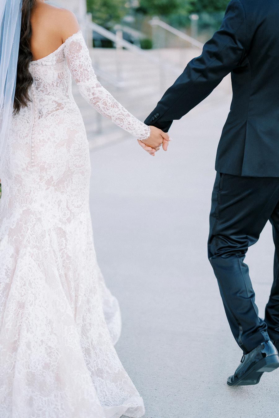 Bride in a lace wedding gown holding hands with groom in formal suit walking outdoors.