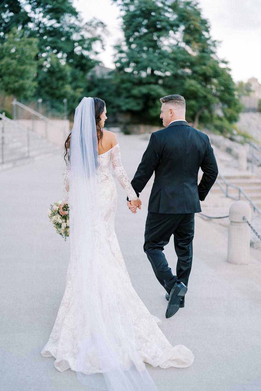 A bride and groom holding hands and walking outdoors, with the bride wearing a white lace gown and veil, and the groom in a black suit, surrounded by lush green trees in a serene setting.