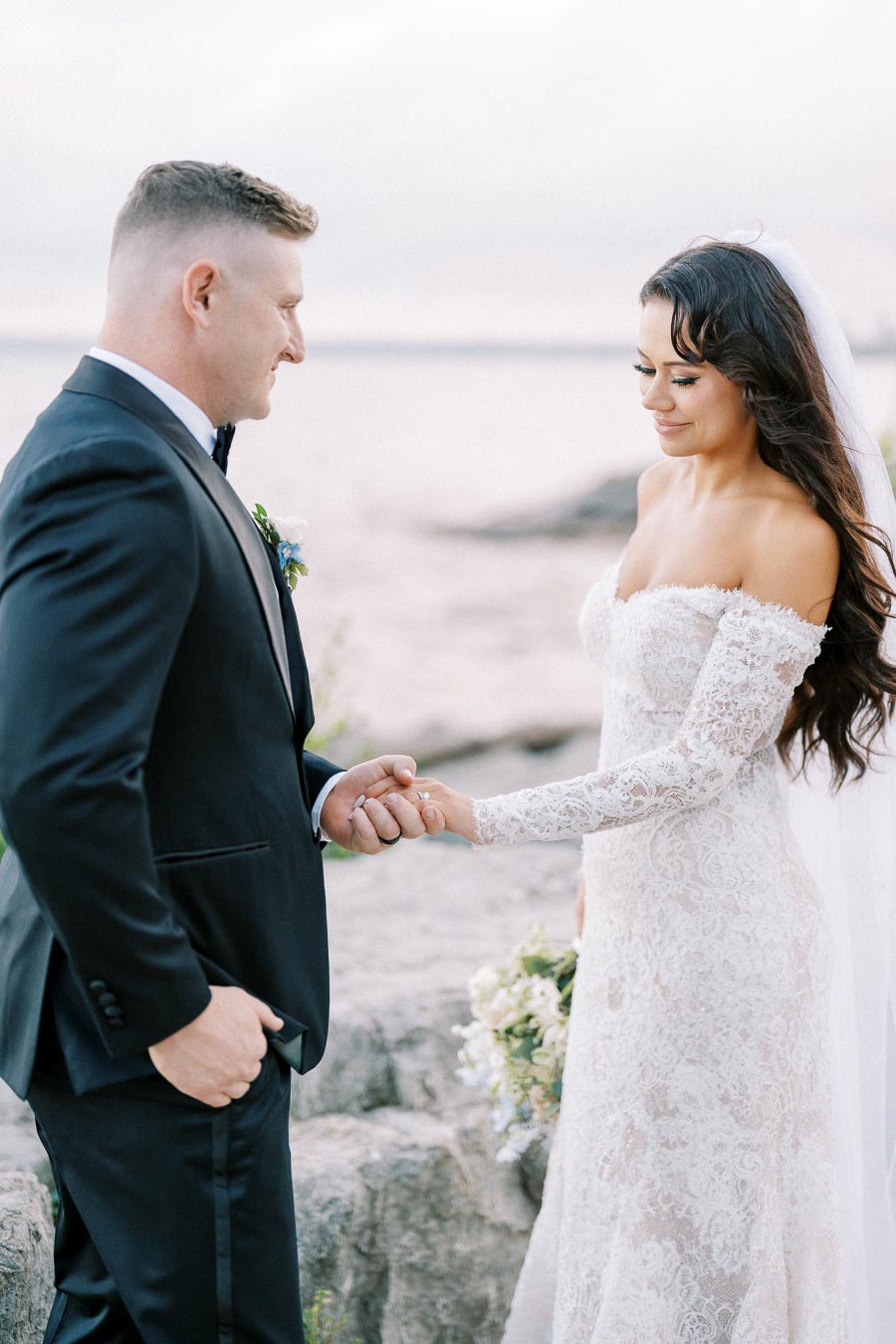 Elegant bride and groom holding hands during outdoor wedding ceremony by the water, bride in lace gown and groom in black tuxedo, picturesque background with soft lighting.