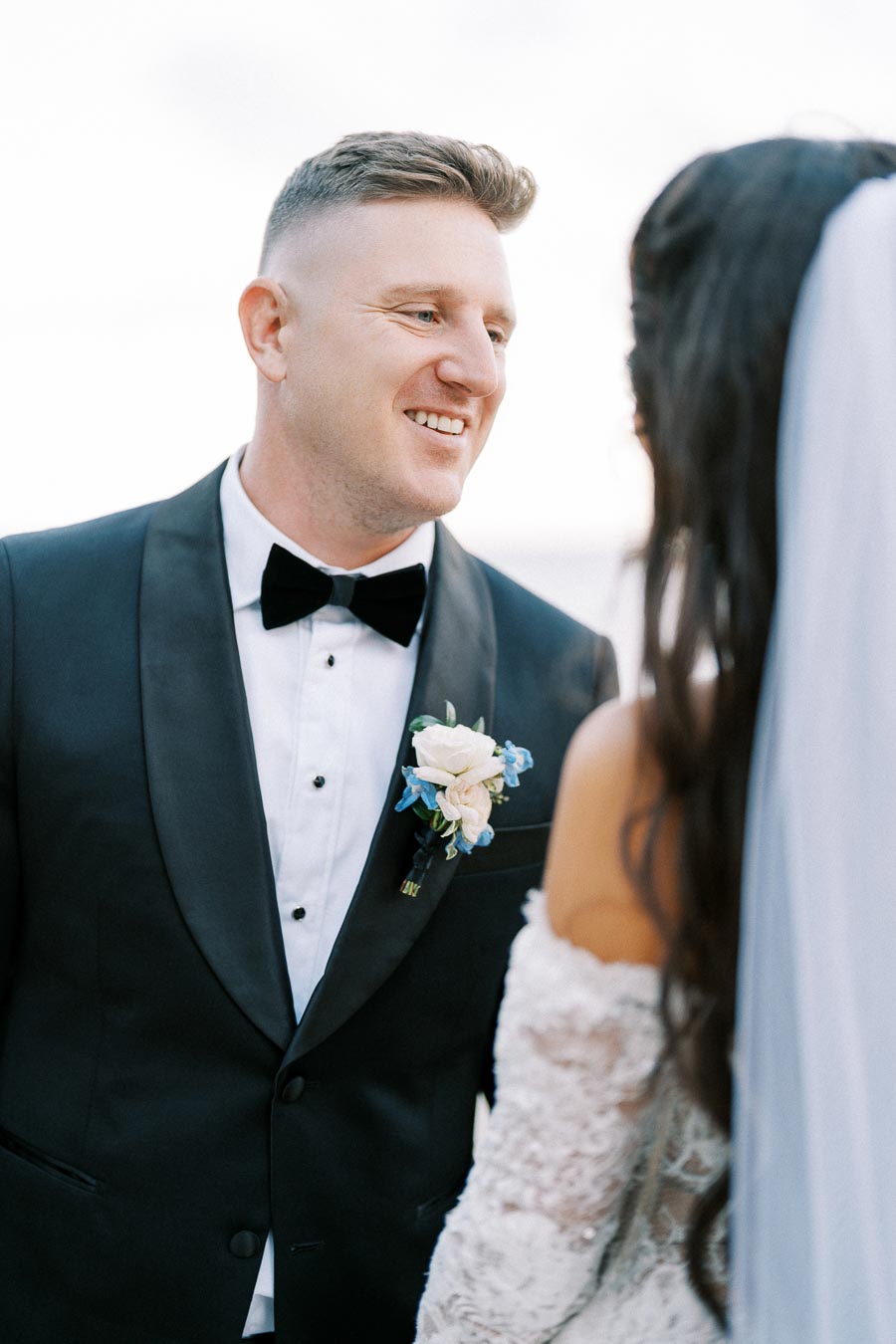 Elegant groom smiling at bride during outdoor wedding ceremony, wearing a classic black tuxedo with a floral boutonnière.