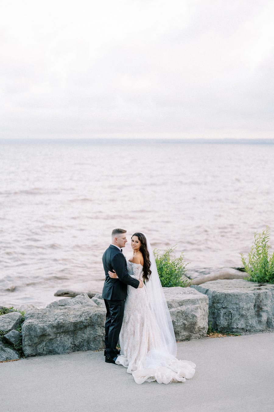 Bride and groom embracing by the waterfront on their wedding day, with scenic lake view and rocky shore in the background