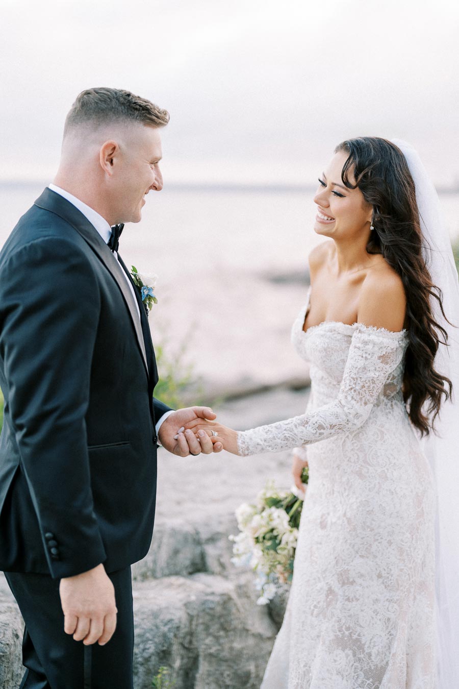 Elegant bride in lace wedding dress smiles at groom in black tuxedo during outdoor ceremony by the water.
