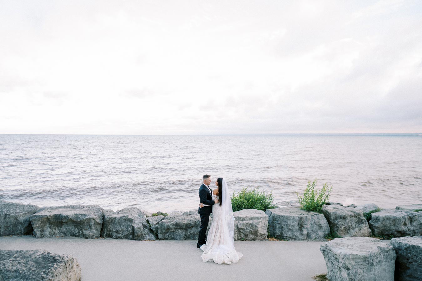 A bride and groom embrace on a rocky shoreline with a vast ocean view under a cloudy sky.